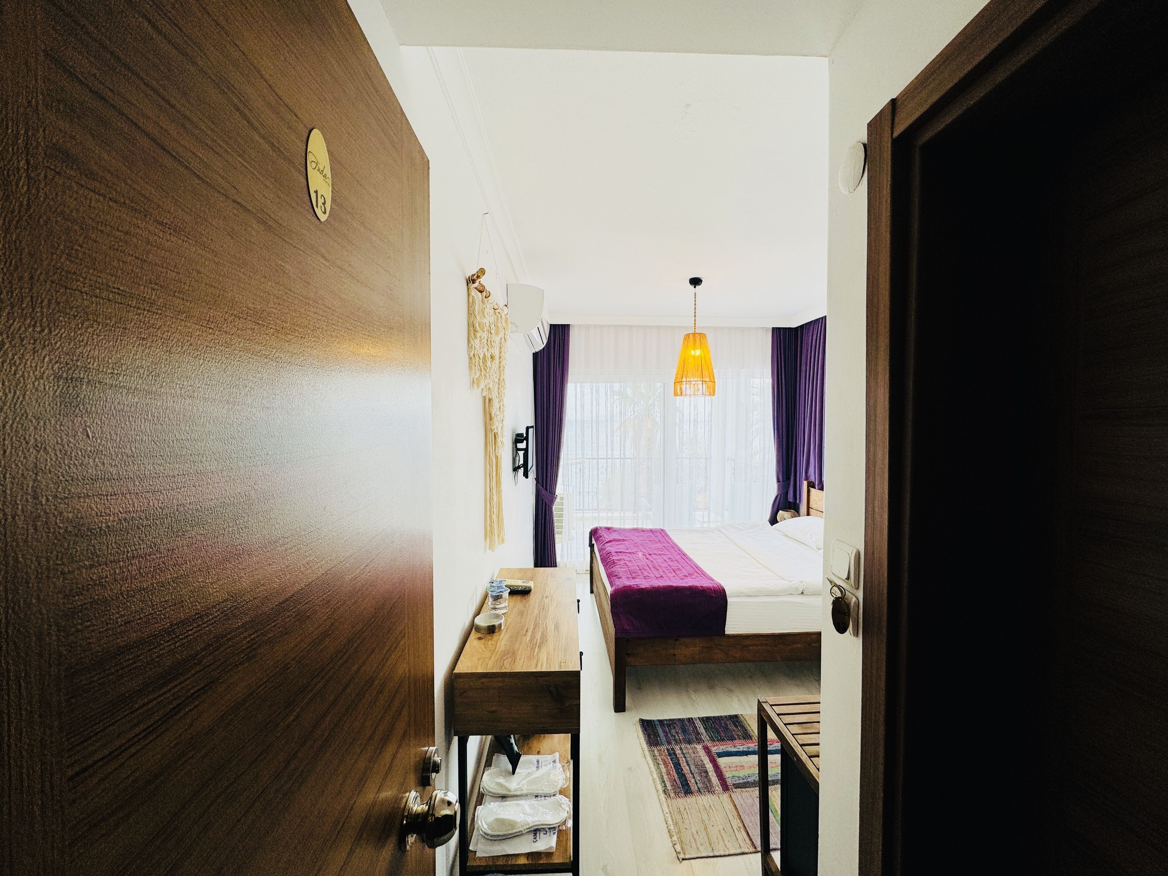 A compact hotel room with a wooden door, a bed with a purple bedspread, a small wooden table, a hanging light fixture, and a view of the street through the window.
