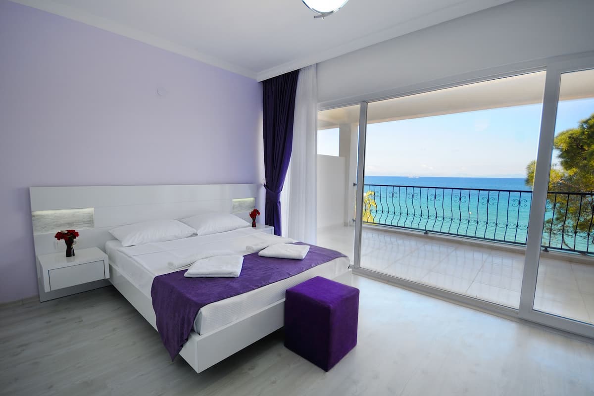 A modern bedroom with a large bed, a nightstand, a purple ottoman, and a balcony with a view of the sea.