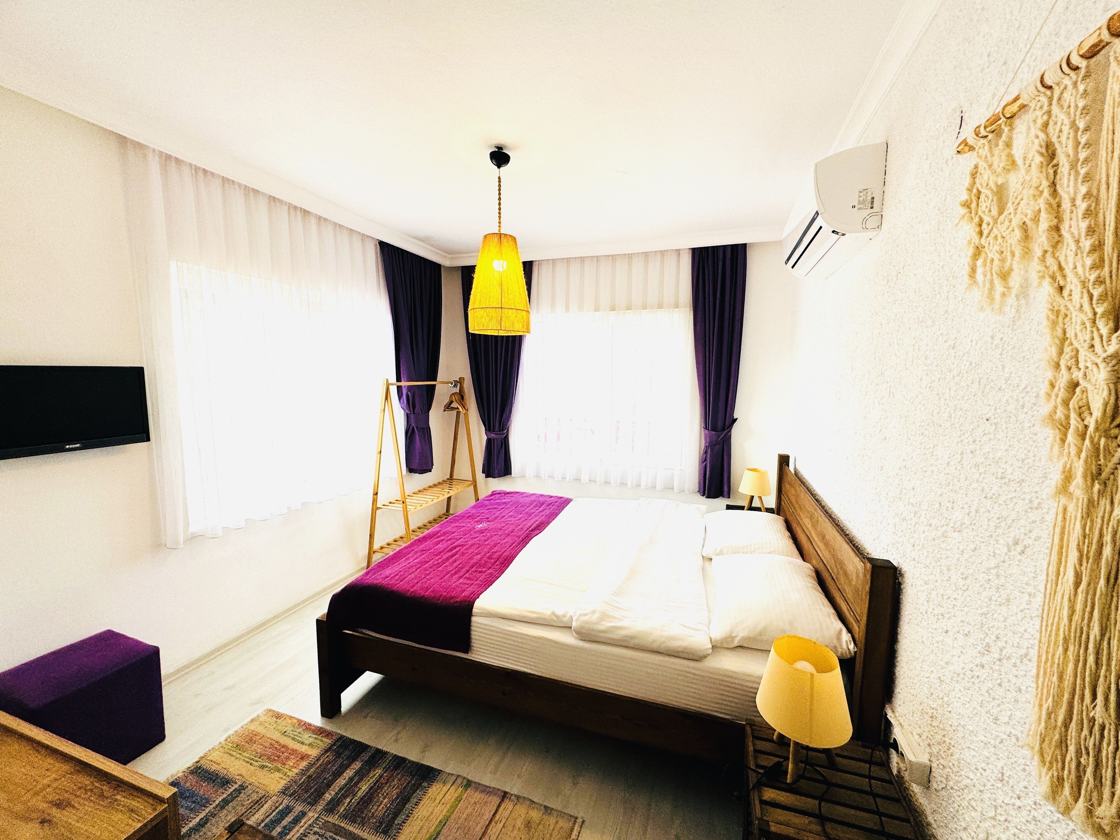 A neatly arranged hotel room with a bed, a wooden ladder, a television mounted on the wall, and a window with curtains.