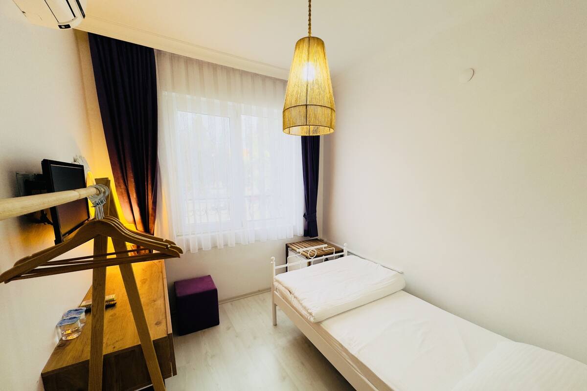 A small, well-lit room with a single bed, a wooden dresser, a hanging light fixture, and a window with curtains.