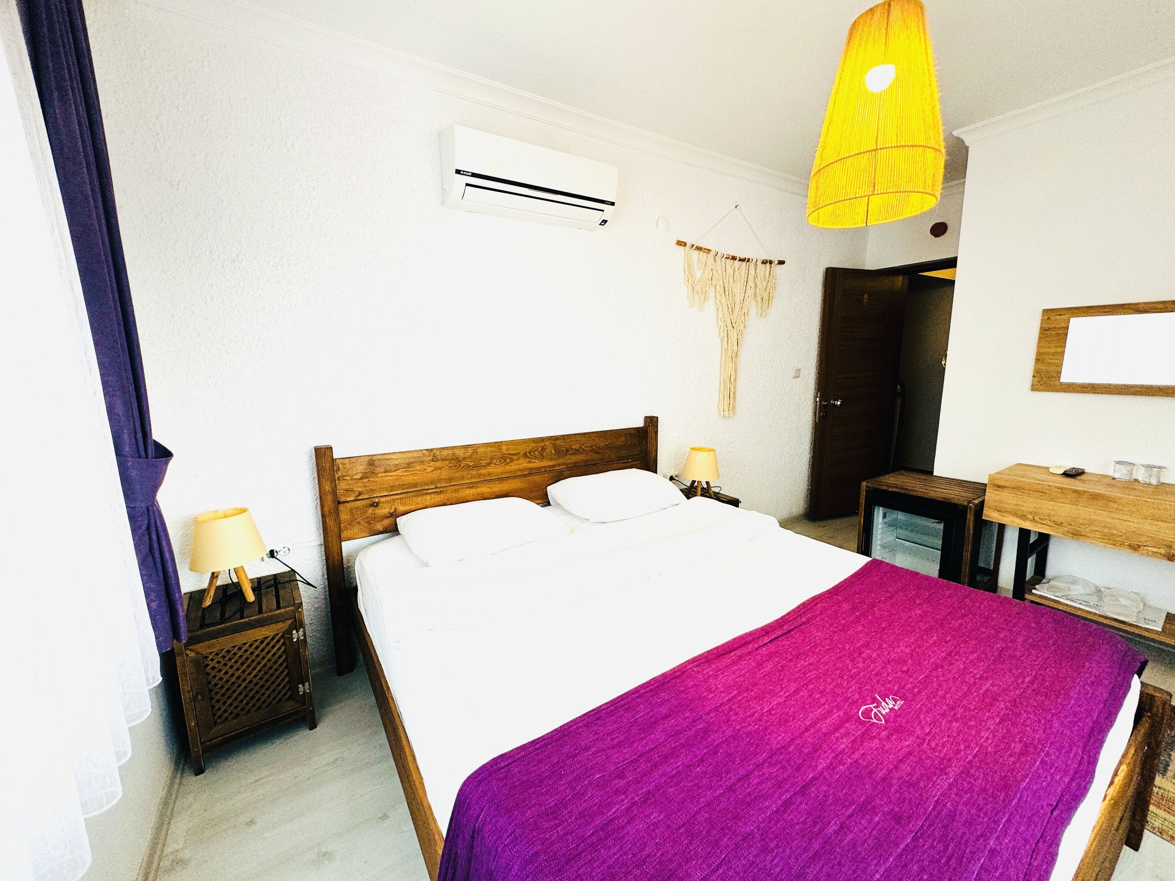 A bedroom with a wooden bed, white bedding, a purple throw, a bedside table with a lamp, an air conditioning unit, a mirror, and a hanging light fixture.