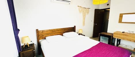 A bedroom with a wooden bed, white bedding, a purple throw, a bedside table with a lamp, an air conditioning unit, a mirror, and a hanging light fixture.