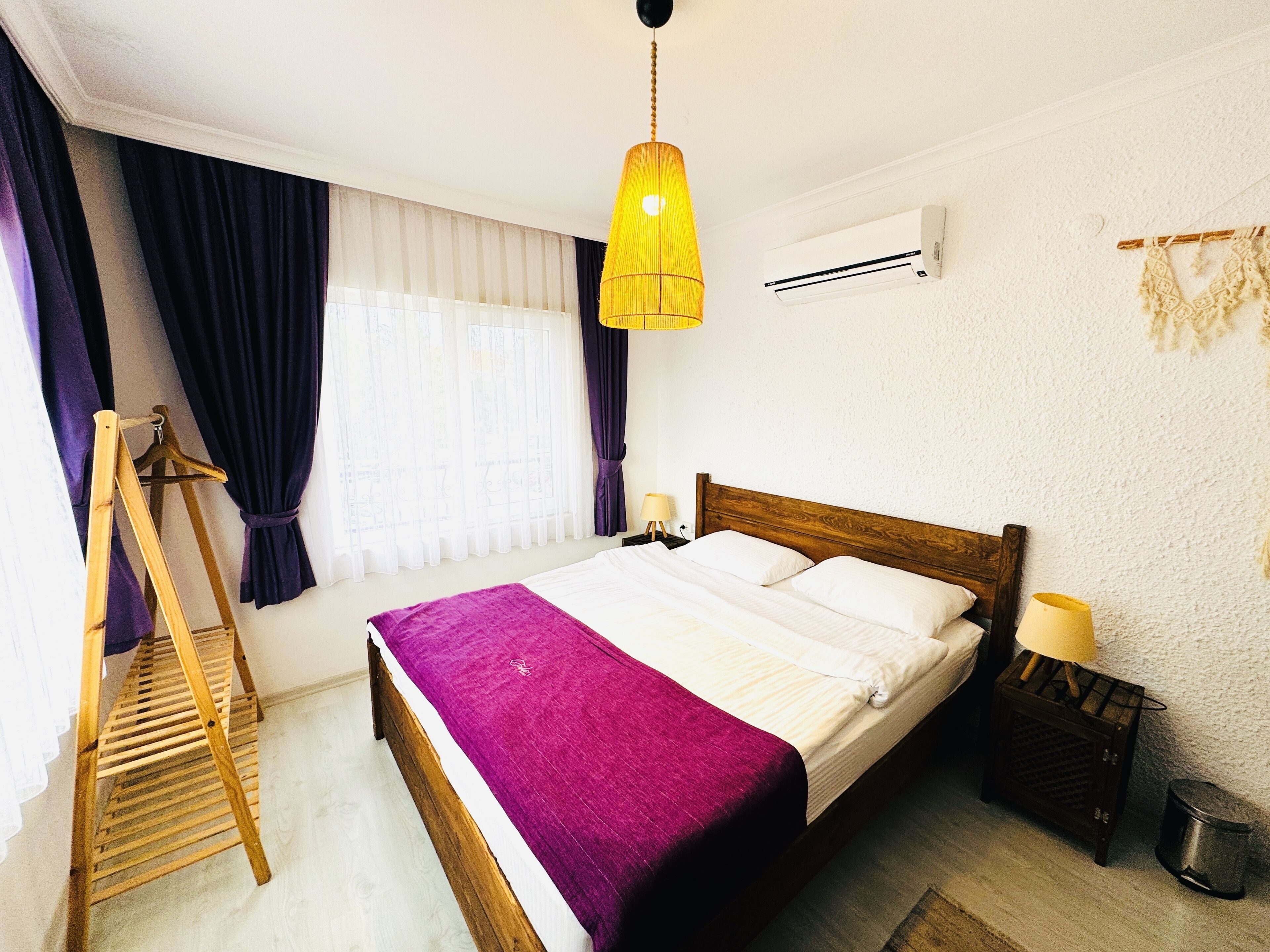 A hotel room with a wooden bed, a purple bedspread, a wooden ladder, a yellow hanging lamp, an air conditioning unit, and a small wooden nightstand with a lamp.