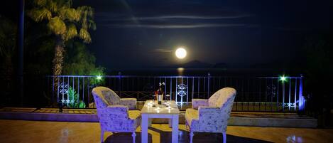 Un área de estar al aire libre con una mesa y sillas, una barandilla con luces decorativas, y una vista de la luna y las montañas al fondo.