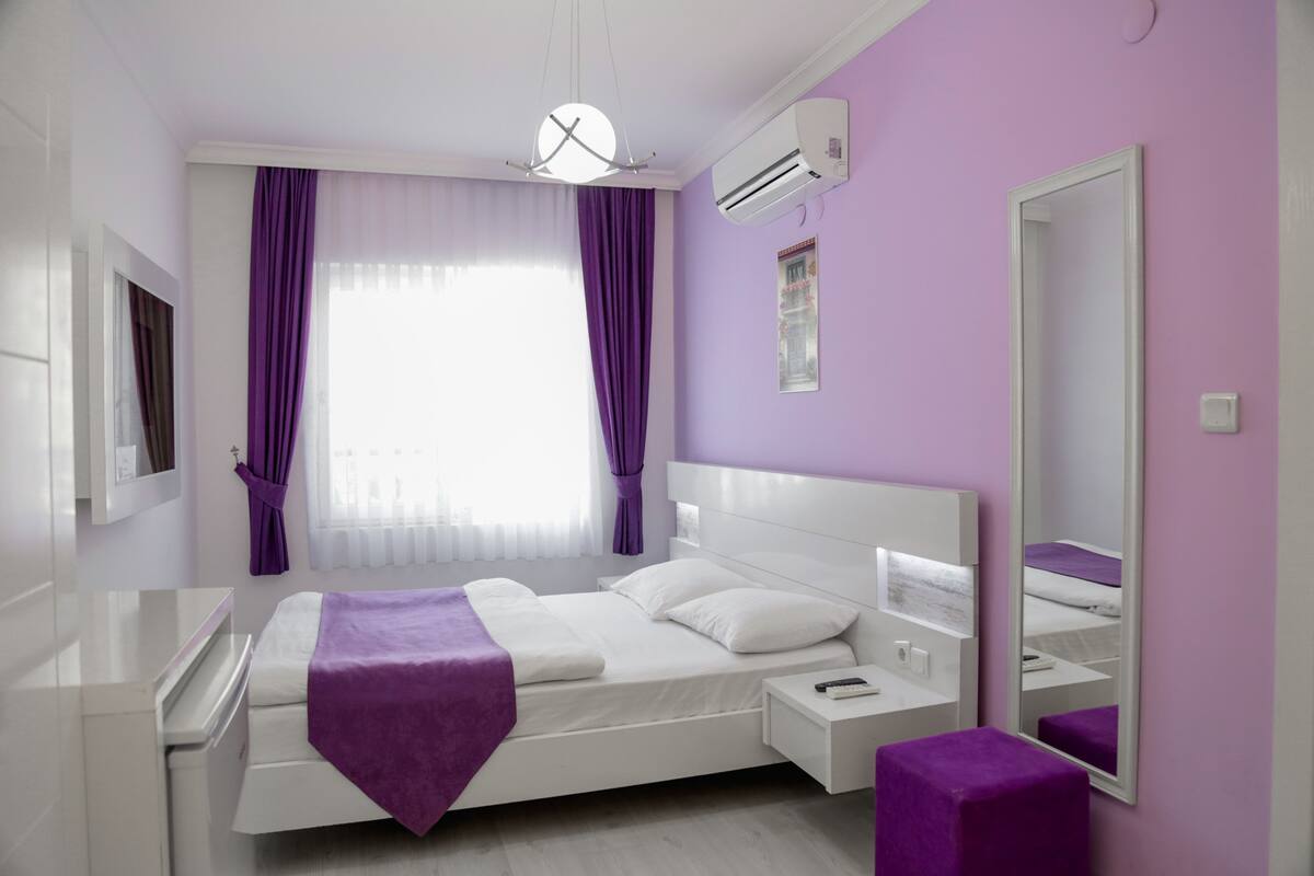 A modern bedroom with a white bed, purple accents, a large mirror, and a window with curtains.