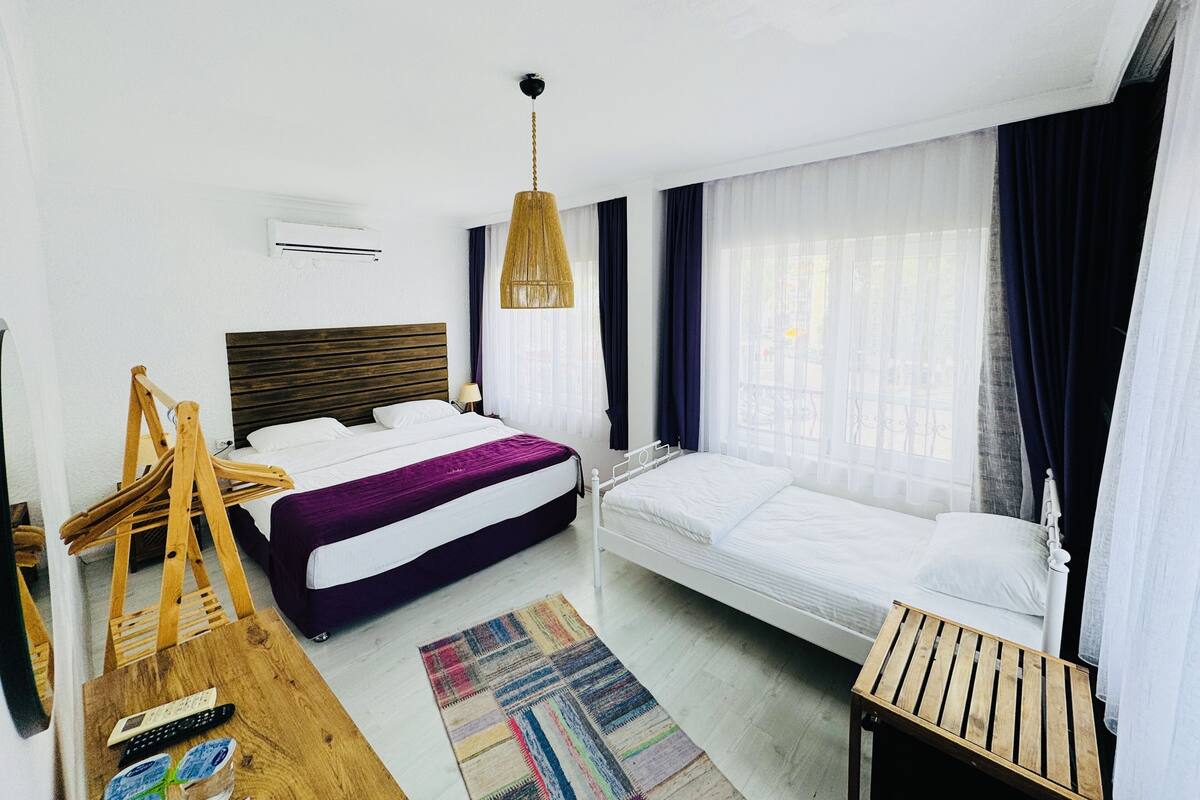 A hotel room with a bed, a smaller bed, a wooden table, a chair, a hanging light fixture, and a window with curtains.