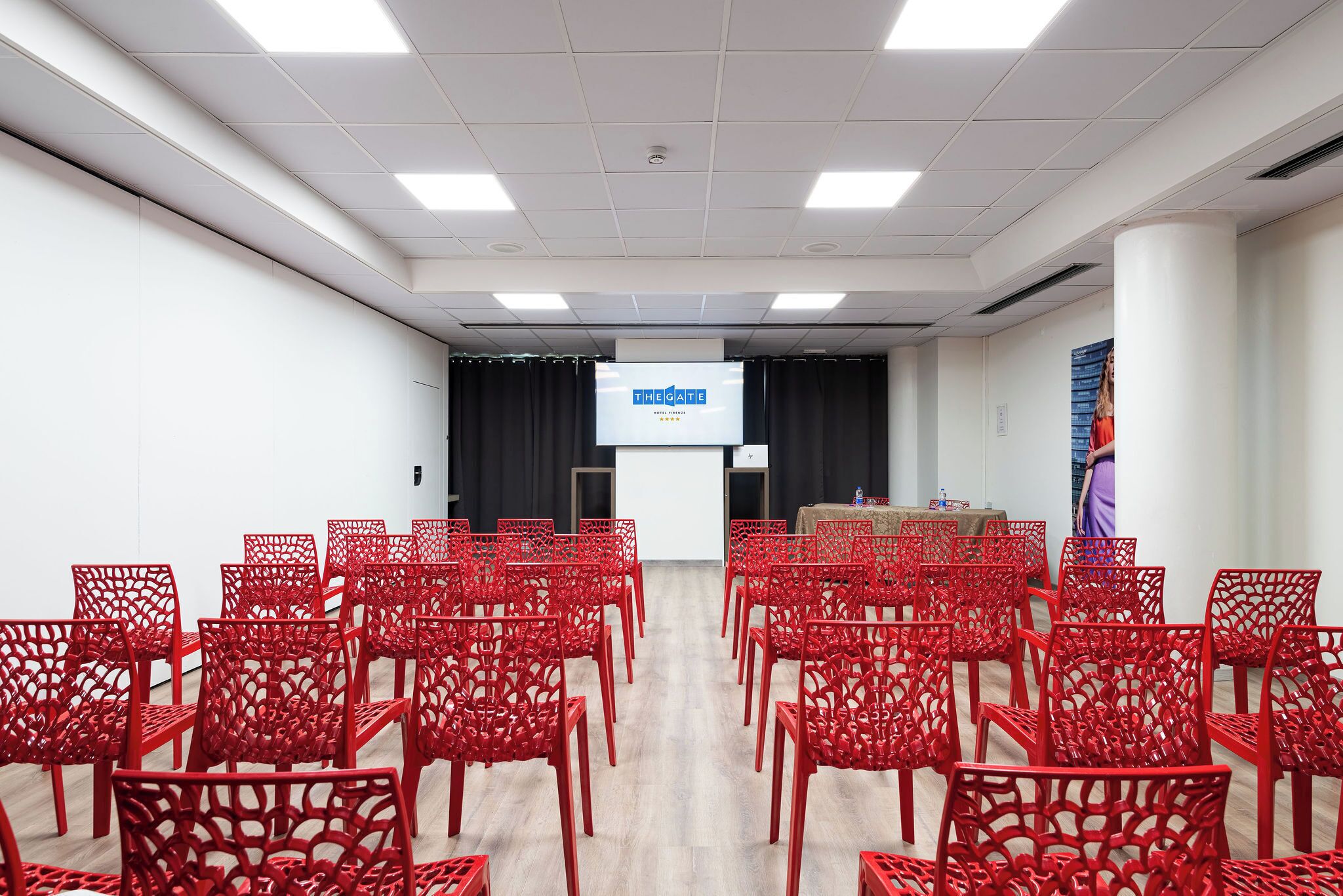 A conference room with red chairs arranged in rows, a projection screen at the front, and a black curtain backdrop.