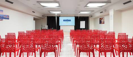 A conference room with rows of red chairs, a large screen displaying 'THE GATE HOTEL FIENZE', and a whiteboard.