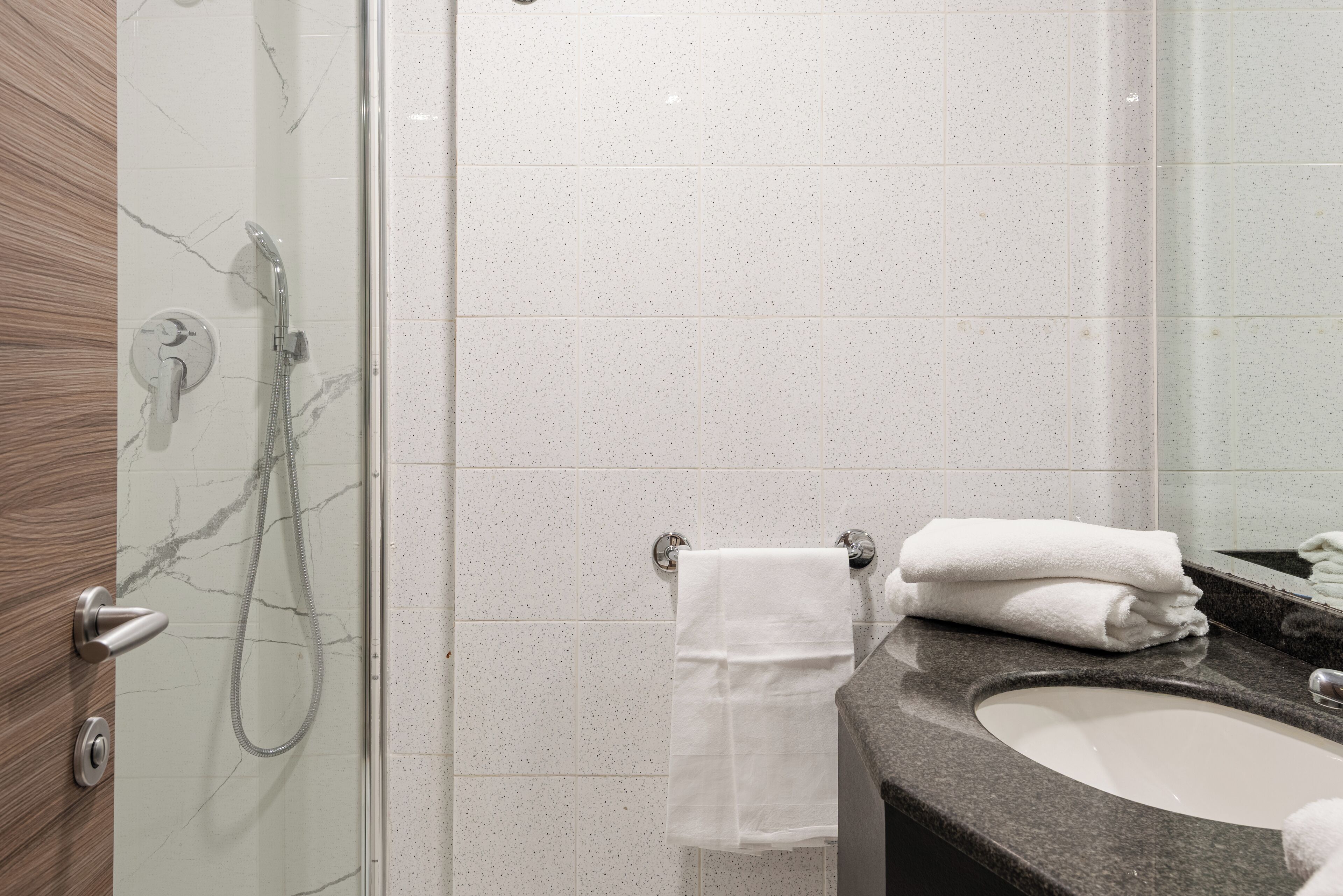 A bathroom with a glass-enclosed shower, a marble-tiled wall, a sink with a dark countertop, and a towel rack with folded towels.