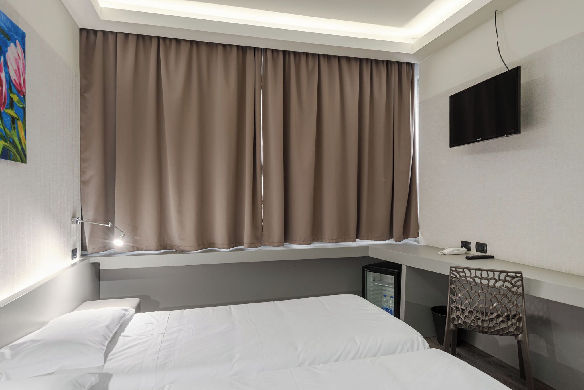 A hotel room with a bed, a desk with a flat-screen TV, a chair, and a window with curtains.