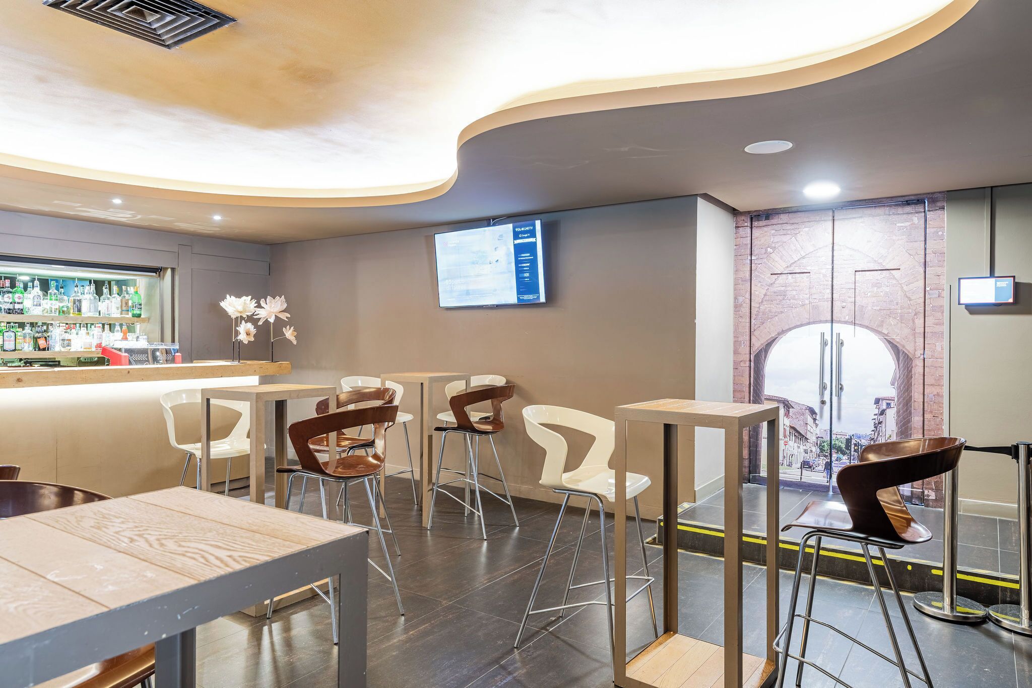 Un espace bar moderne avec un comptoir où sont exposées différentes bouteilles, plusieurs tabourets et un écran fixé au mur.