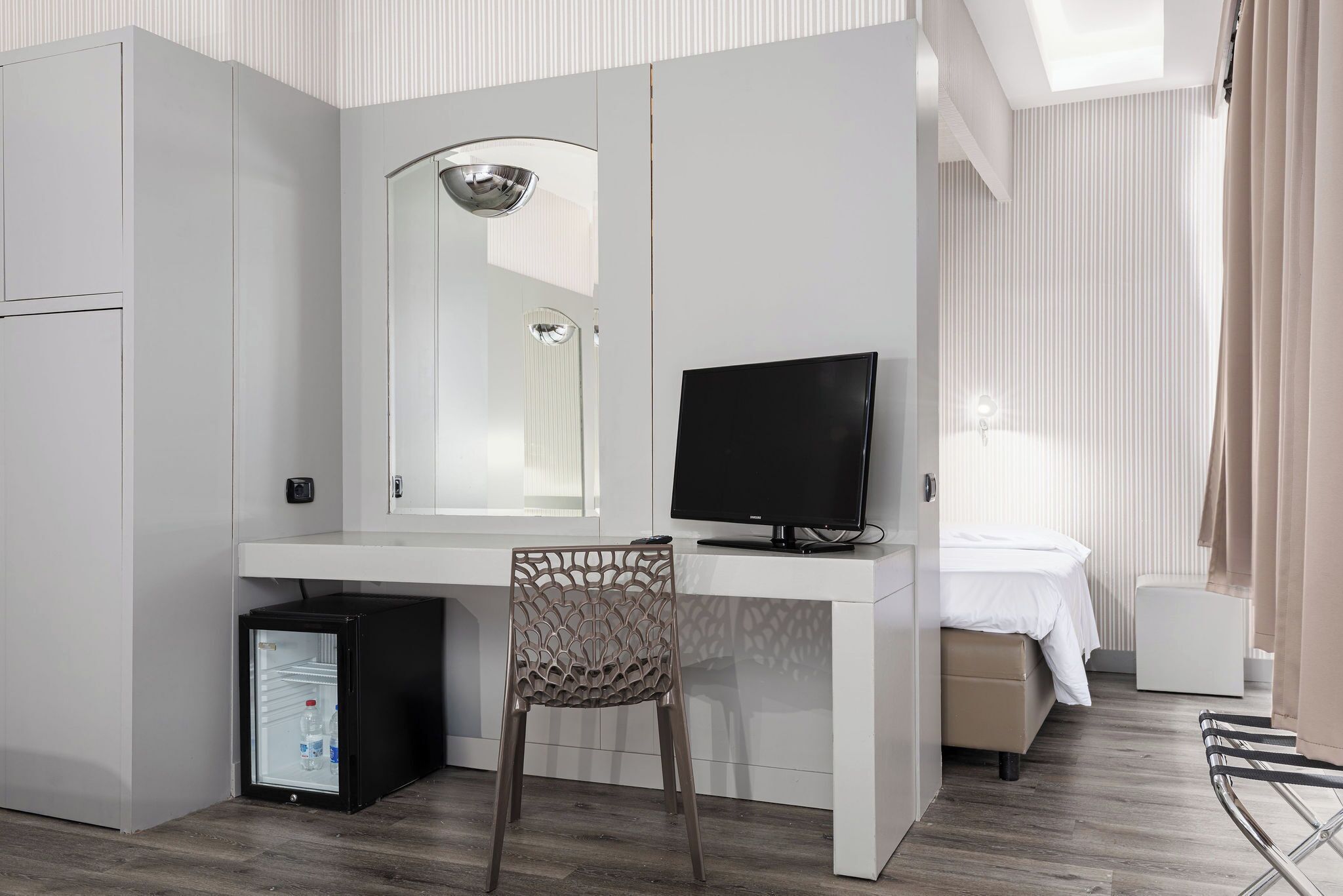Ein kompaktes Hotelzimmer mit weißem Schreibtisch, Fernseher, kleinem Kühlschrank und einem Bett mit weißer Bettdecke.