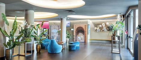 A spacious lobby with modern furniture, large potted plants, and a view of the city through a glass wall.