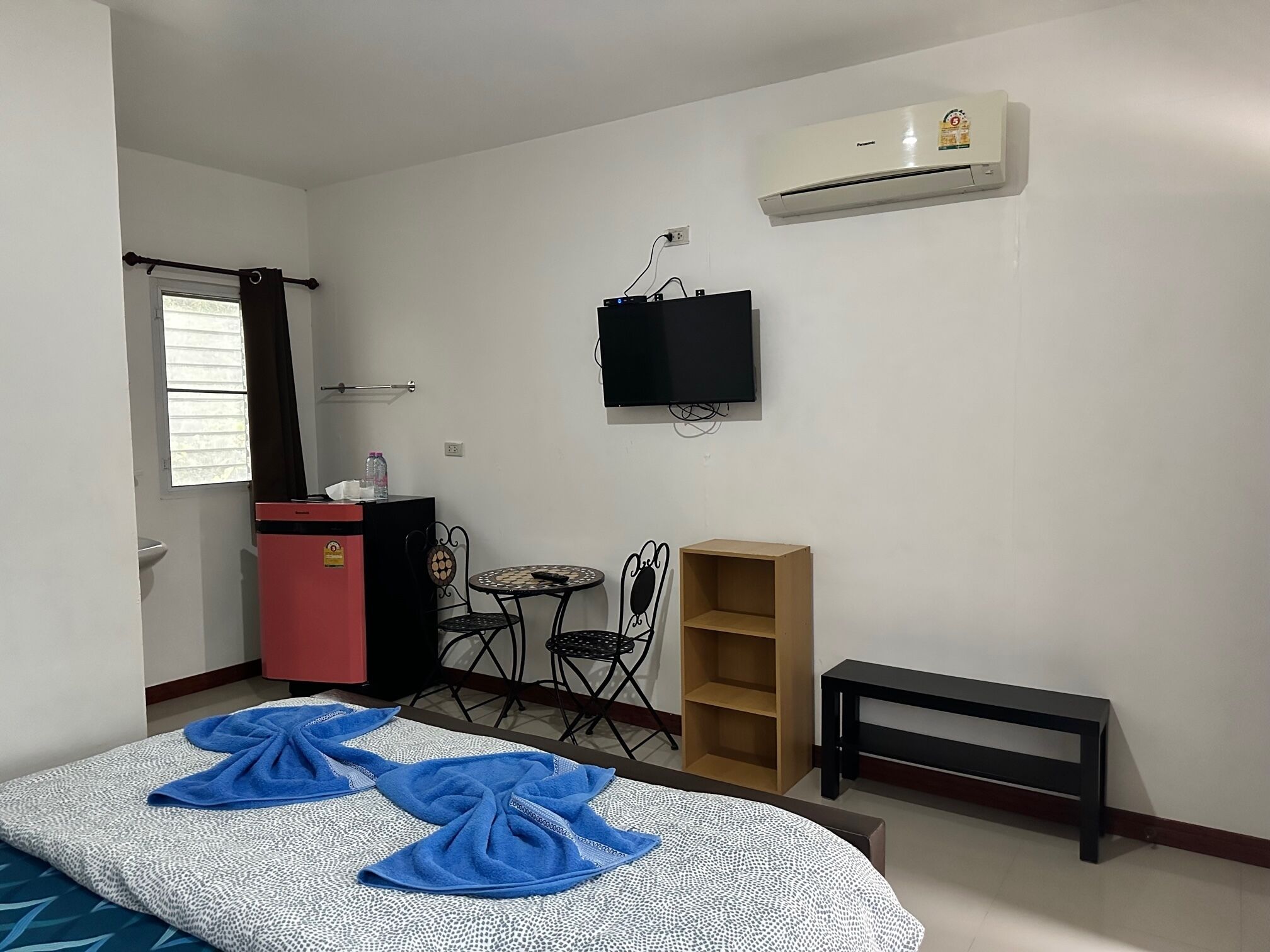 A small, tidy room with a bed, a desk, a chair, a TV, and an air conditioning unit.