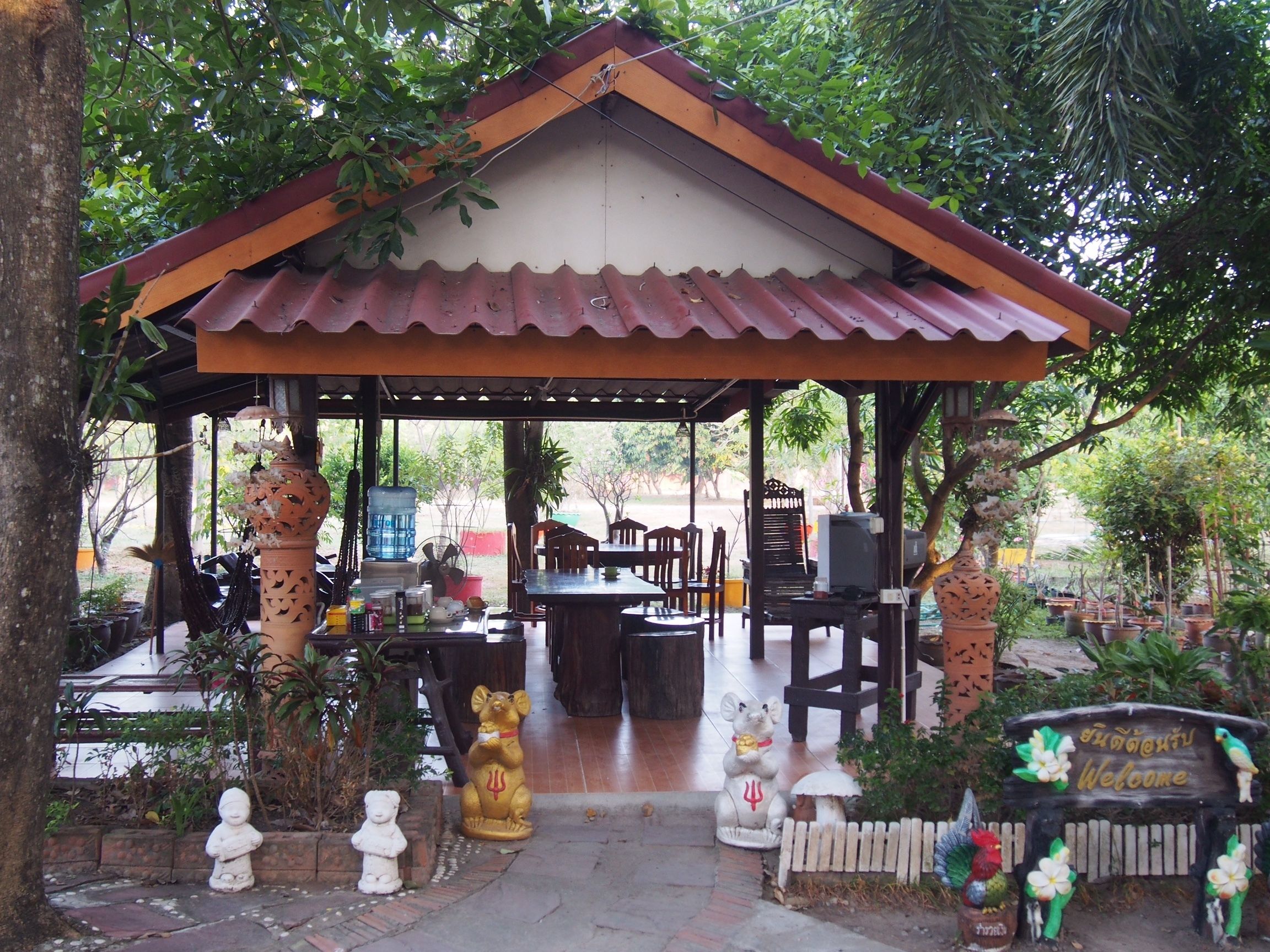 A covered outdoor seating area with tables and chairs, surrounded by potted plants and decorative statues.