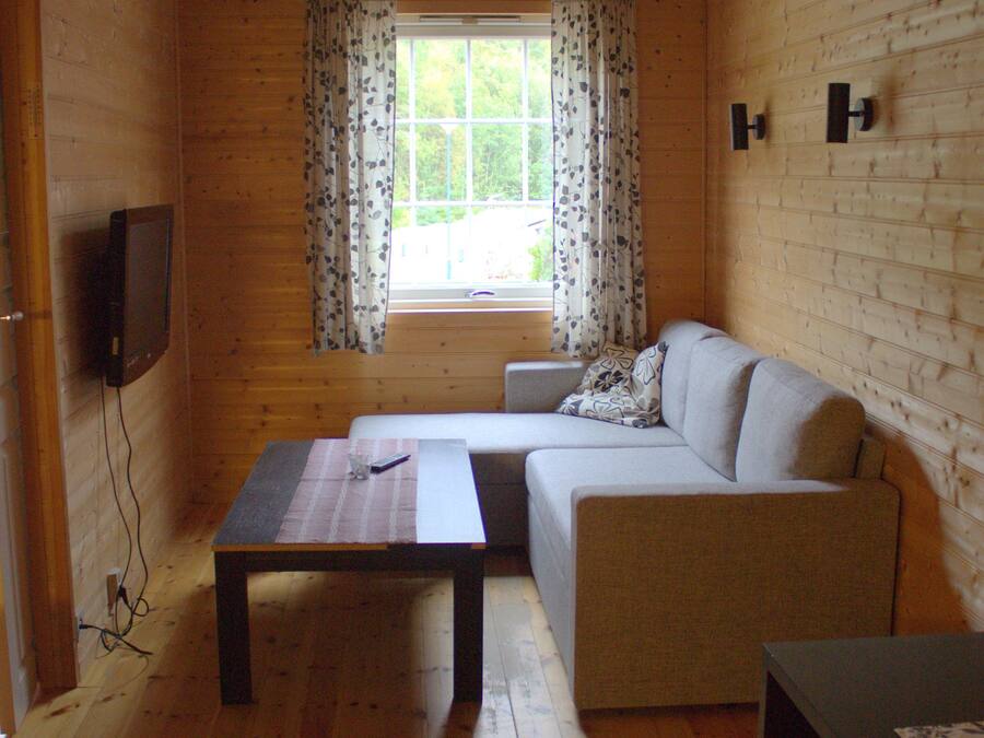 A wooden cabin room with a sofa, a table, a TV, and a window with curtains.