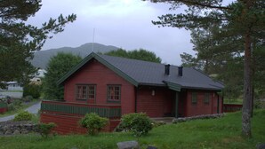 A red wooden cabin with a green roof, surrounded by a stone wall, and a stone pathway leading to the entrance. - Sjøholt Camping (Ålesund)