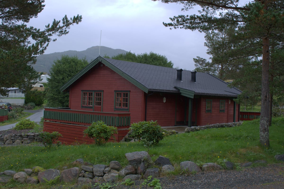 A red wooden cabin with a green roof, surrounded by a stone wall, and a stone pathway leading to the entrance.