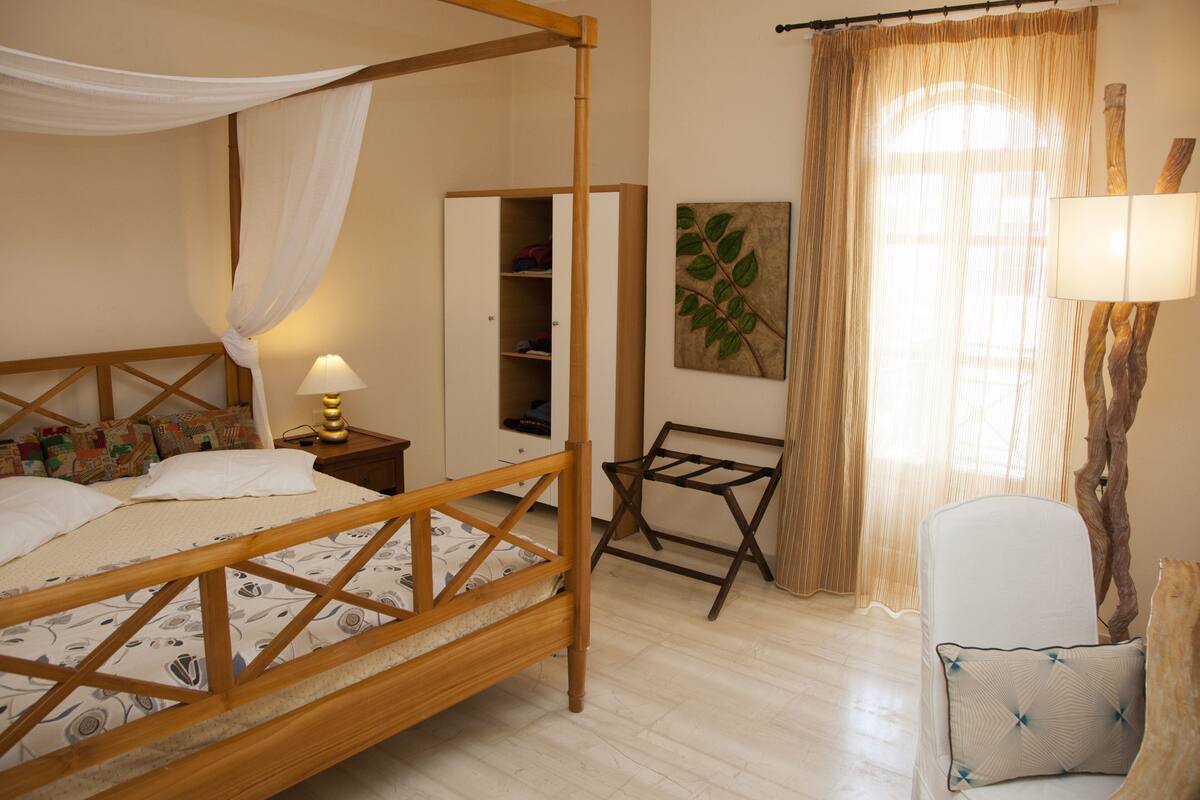 Ein Schlafzimmer mit Himmelbett, Nachttisch mit Lampe, Kleiderschrank, Stuhl und Fenster mit durchsichtigen Vorhängen.