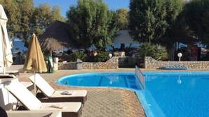 A swimming pool with lounge chairs and umbrellas, surrounded by trees and a stone wall. - Troulakis Beach Hotel (Chania)