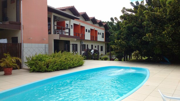 A swimming pool with a tiled deck, surrounded by a building with multiple balconies and a fenced area with potted plants. - Hospedaria Ondas do Rosa (Garopaba)