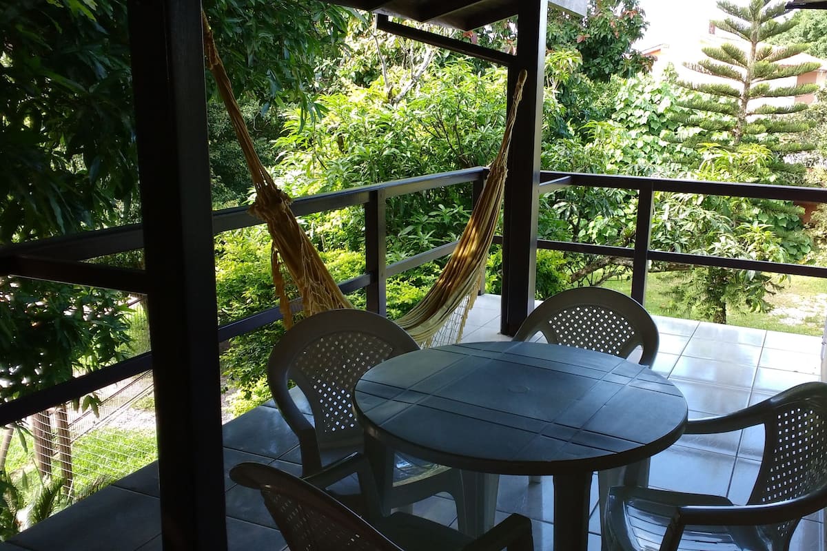 A covered outdoor seating area with a table and chairs, surrounded by lush greenery.