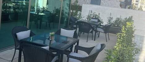 An outdoor seating area with tables and chairs, a glass wall reflecting the building, and potted plants.