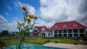 Un edificio grande con techo rojo y varias ventanas, rodeado de un césped cuidado y una piscina.