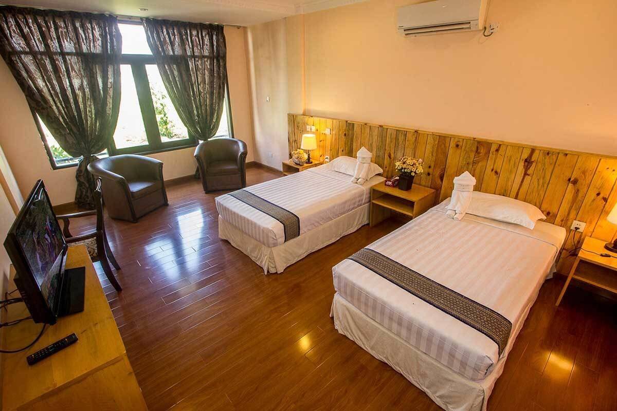 A hotel room with two single beds, a wooden headboard, a TV, a desk, and a window with curtains.