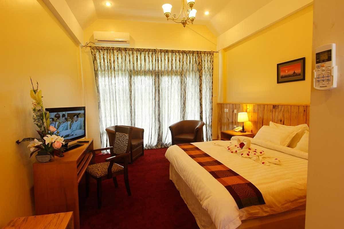 Une chambre d’hôtel comprenant un lit, deux chaises, un bureau avec une télévision, une table de chevet en bois avec une lampe, et une grande fenêtre avec des rideaux.