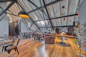 Amplia habitación tipo loft con techos altos, vigas a la vista y techo inclinado. Hay varias zonas de estar con mesas y sillas, y un comedero decorativo en una pared.