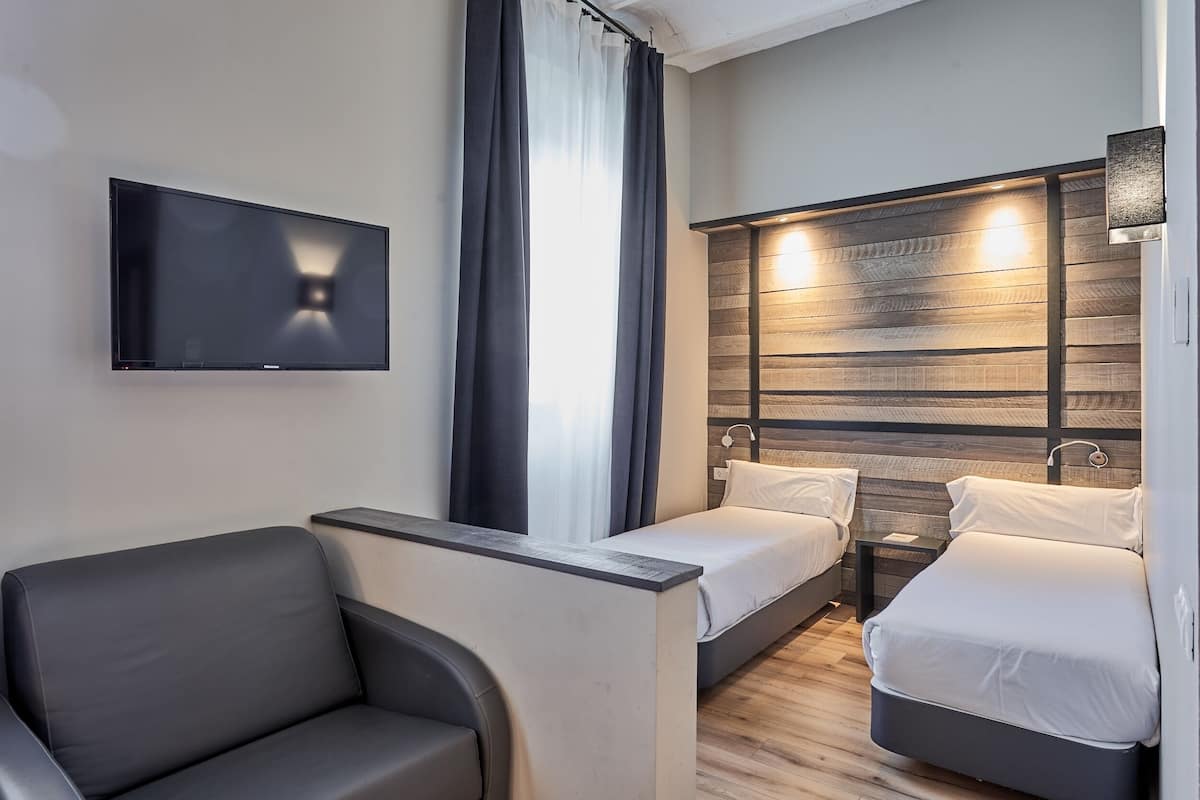 A modern hotel room with a flat-screen TV mounted on the wall, a sofa, two single beds, a nightstand, and a wooden accent wall.