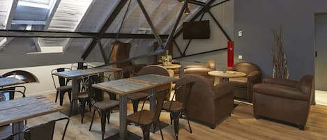 A spacious loft with a high ceiling, exposed beams, and a skylight. There is a dining area with wooden tables and chairs, and a seating area with leather sofas and a large pendant light.