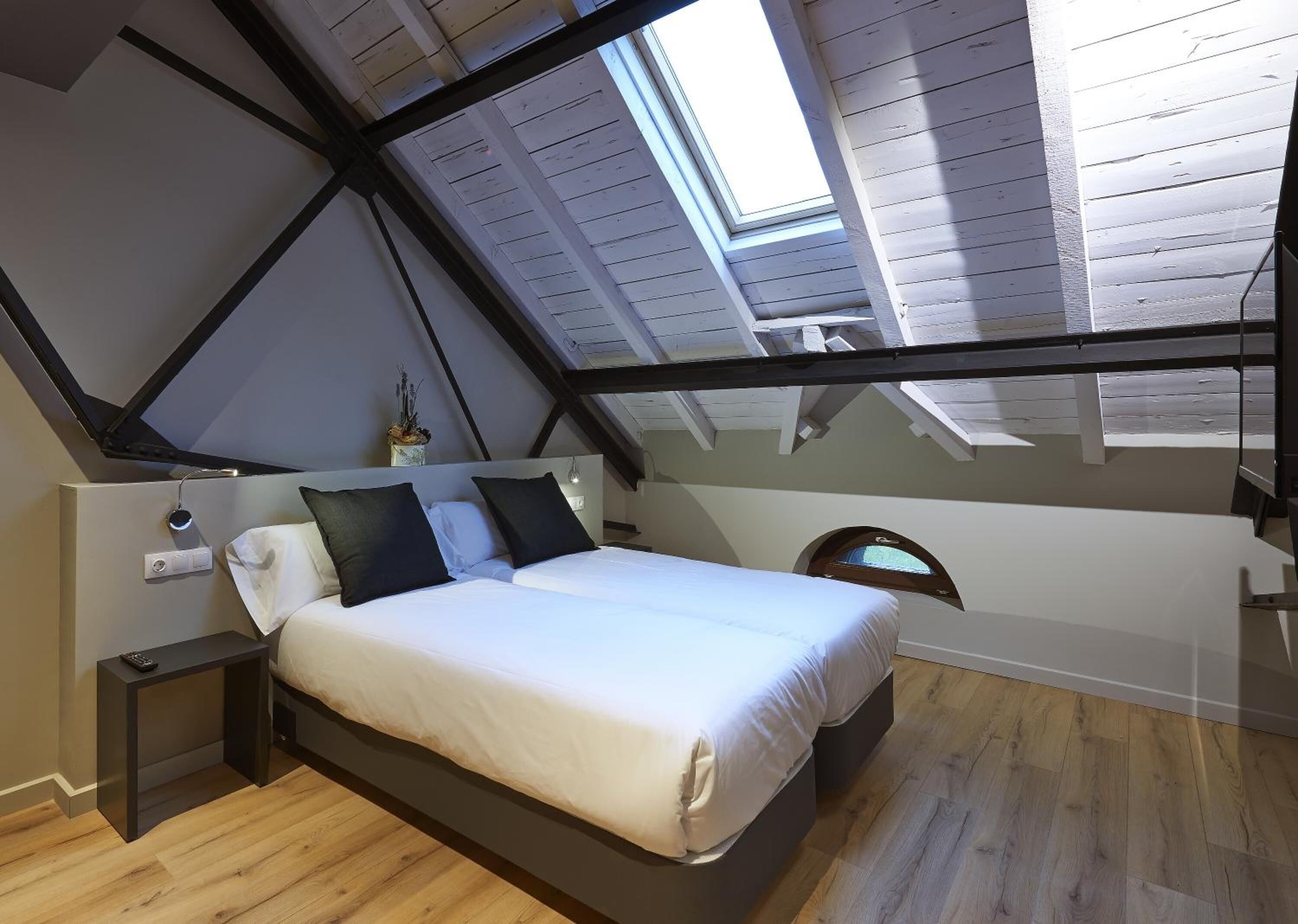 Un dormitorio con techo inclinado, una cama, una mesita de noche y un ventanal pequeño.