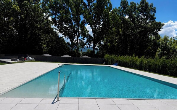 Una piscina rectangular con superficie de agua azul cristalina, rodeada por un borde de baldosas y abundante vegetación.