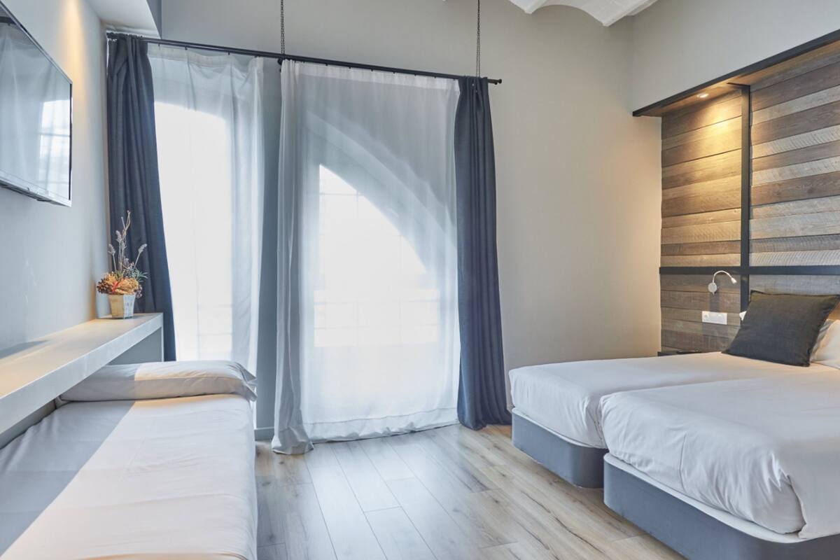 A modern bedroom with two beds, a wooden accent wall, a bedside table with a vase, and sheer curtains allowing natural light.