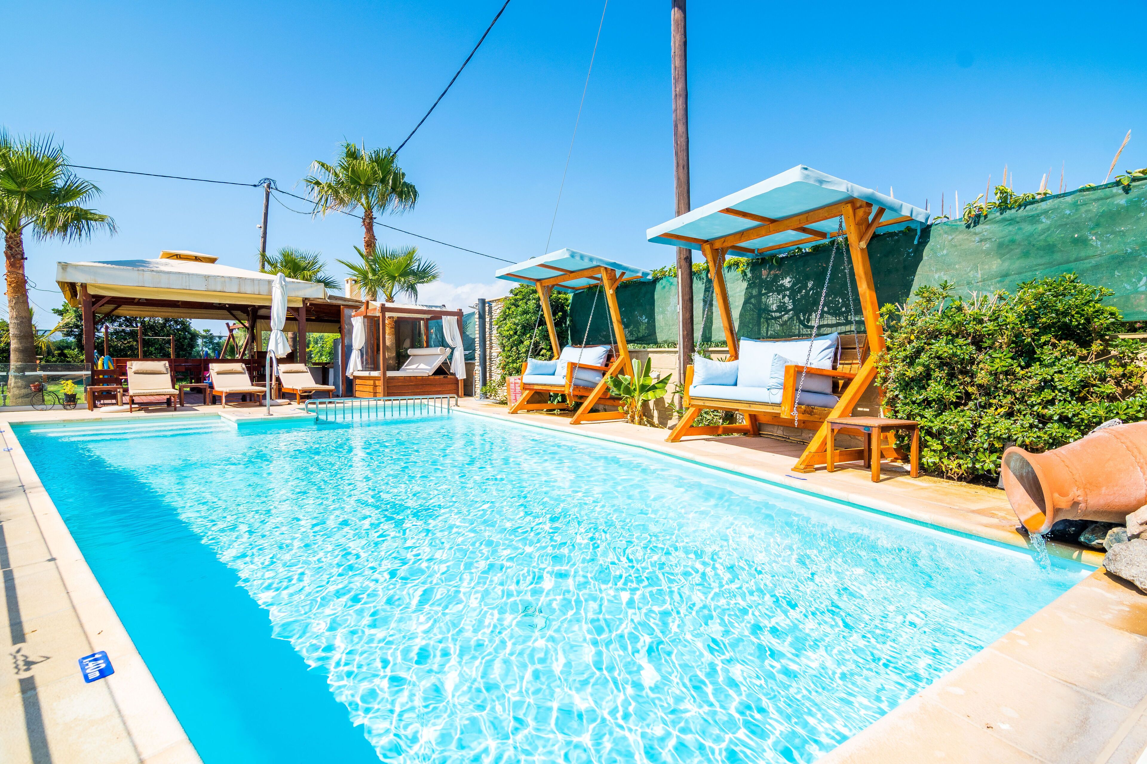 A clear swimming pool with lounge chairs and umbrellas, surrounded by palm trees and a covered seating area.