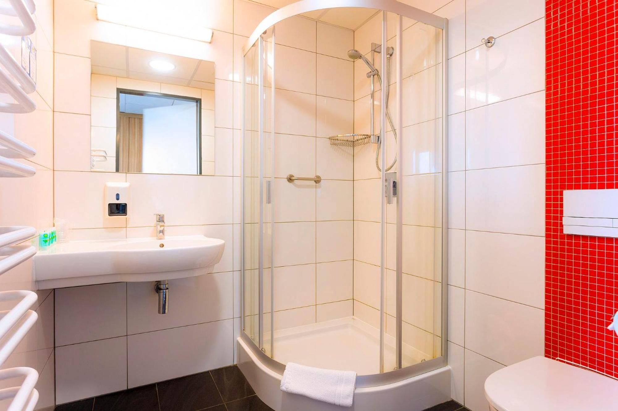 Un baño moderno con ducha de vidrio, un lavamanos blanco con dispensador de jabón y una pared de acento con azulejos rojos.