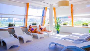 A sunlit indoor relaxation area with white lounge chairs, large windows, and potted plants.