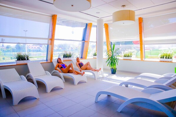 A sunlit indoor relaxation area with white lounge chairs, large windows, and potted plants.