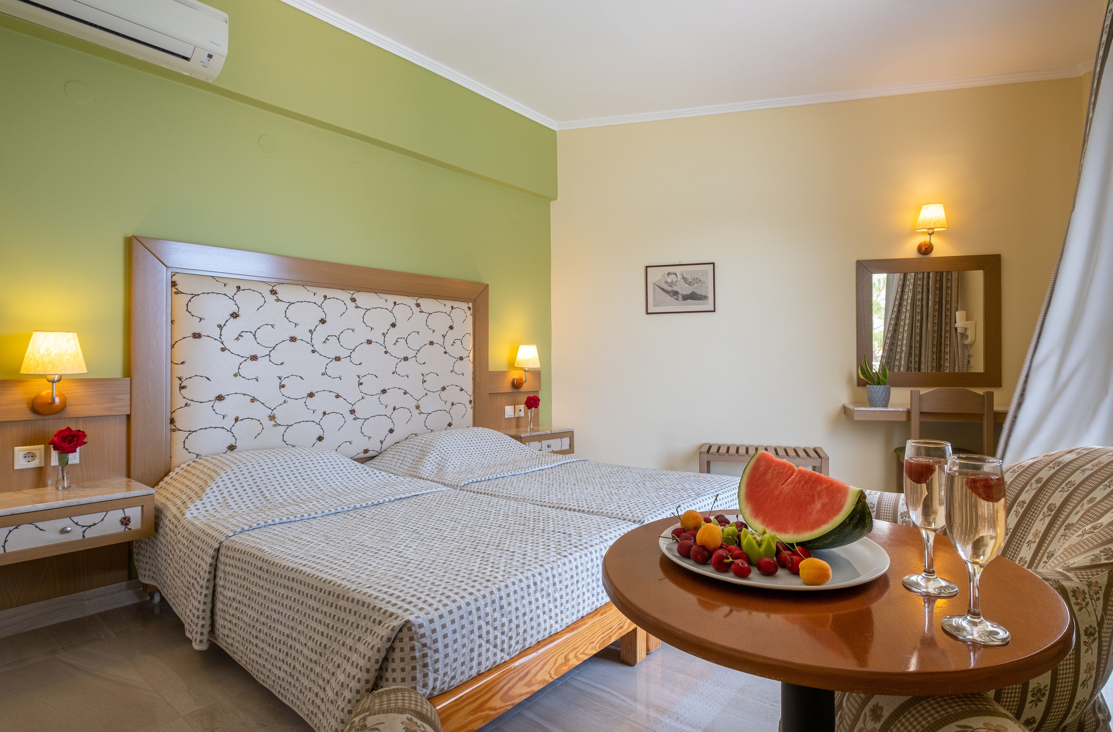 Een hotelkamer met twee bedden, een nachtkastje met een lamp, een ronde tafel met fruit en drankjes, en een spiegel met verlichting.