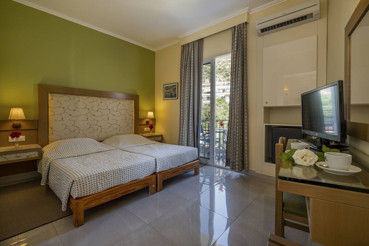 A hotel room with a bed, bedside lamps, a TV, a small table with a cup and saucer, and a view of the outside balcony.