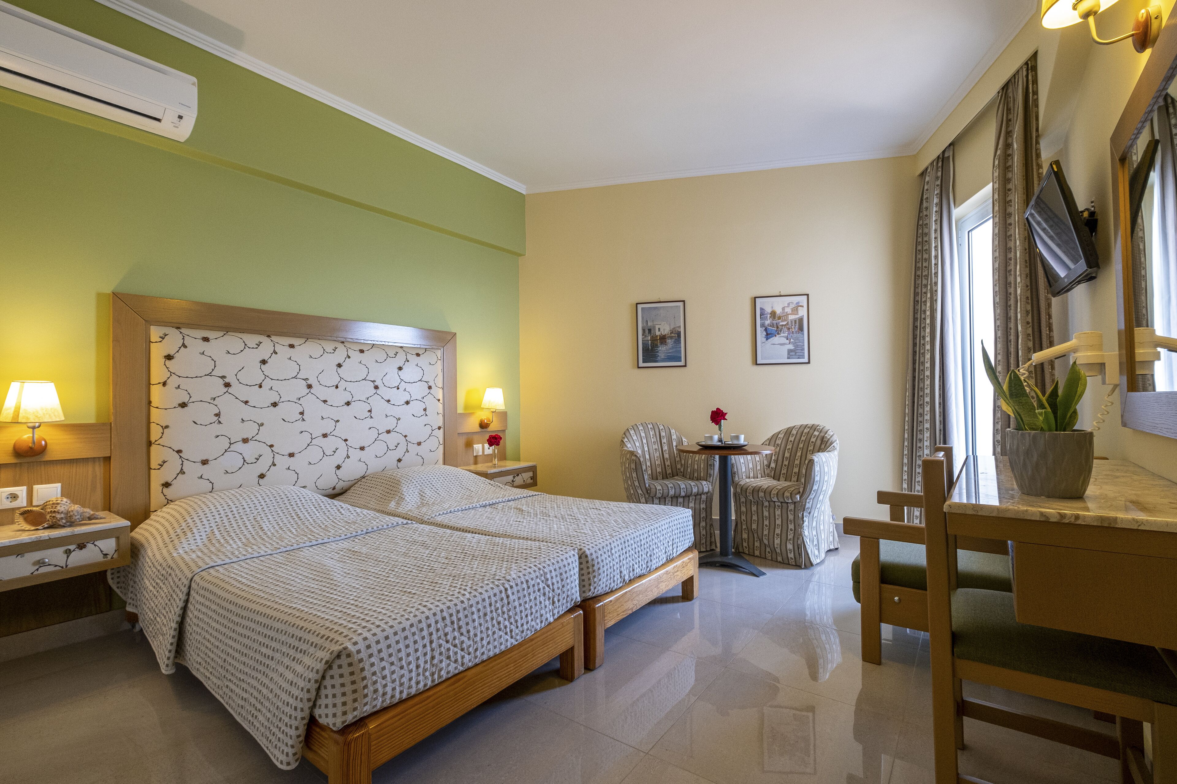A hotel room with two beds, a small table, a chair, a TV, and a potted plant.