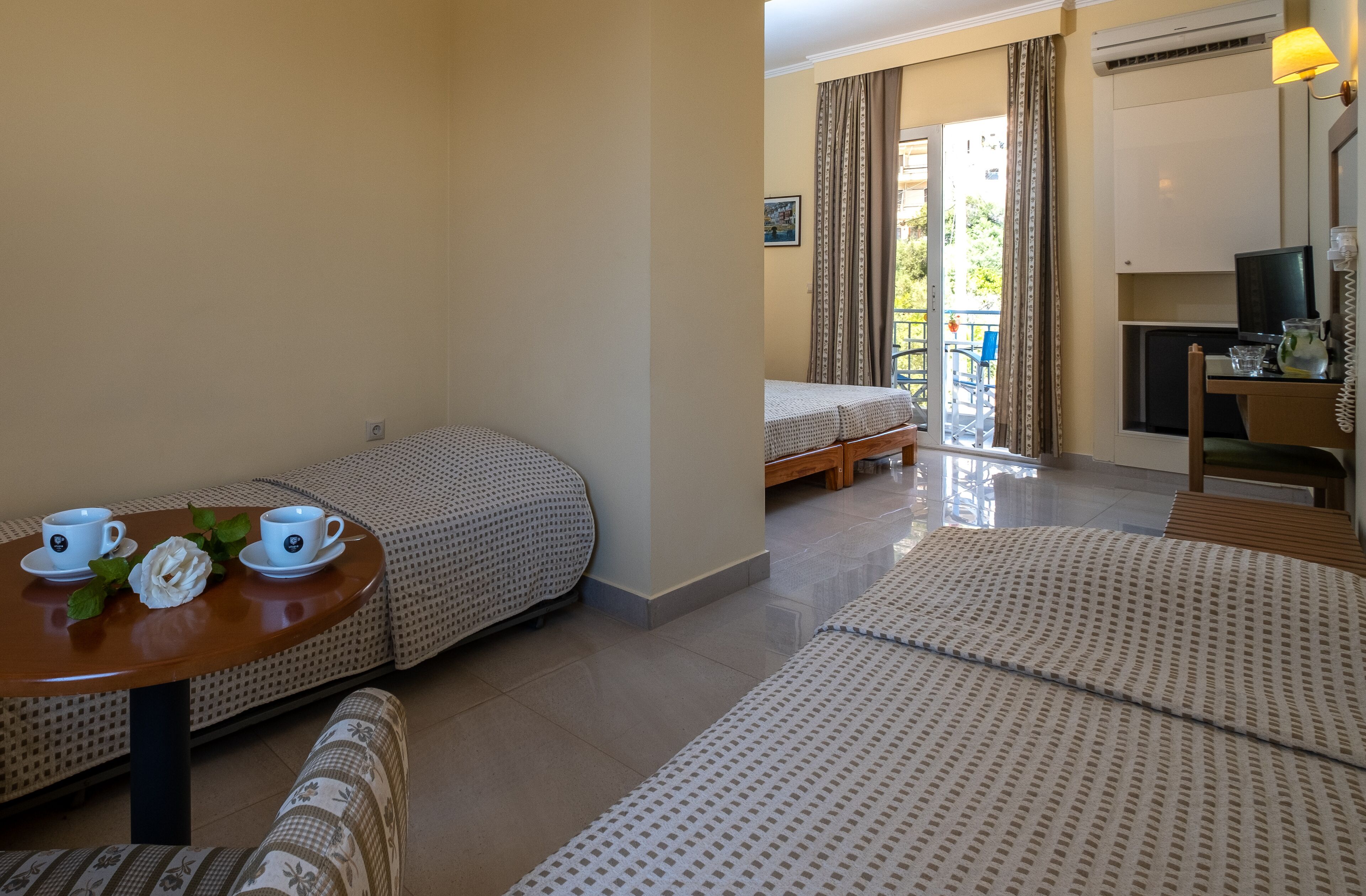 A hotel room with two beds, a small table with cups and a flower, a sofa, a TV, and a balcony with a chair.