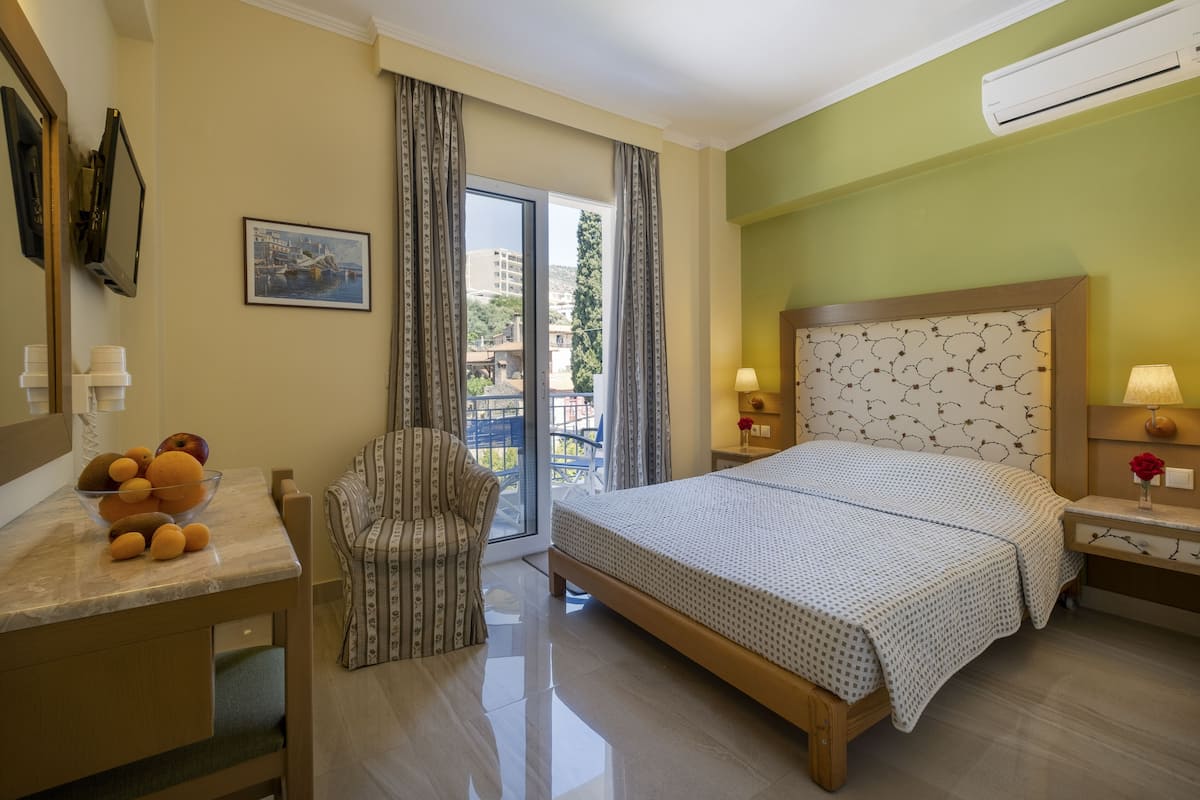 A hotel room with a bed, a desk with a bowl of fruit, a chair, and a balcony with a view.