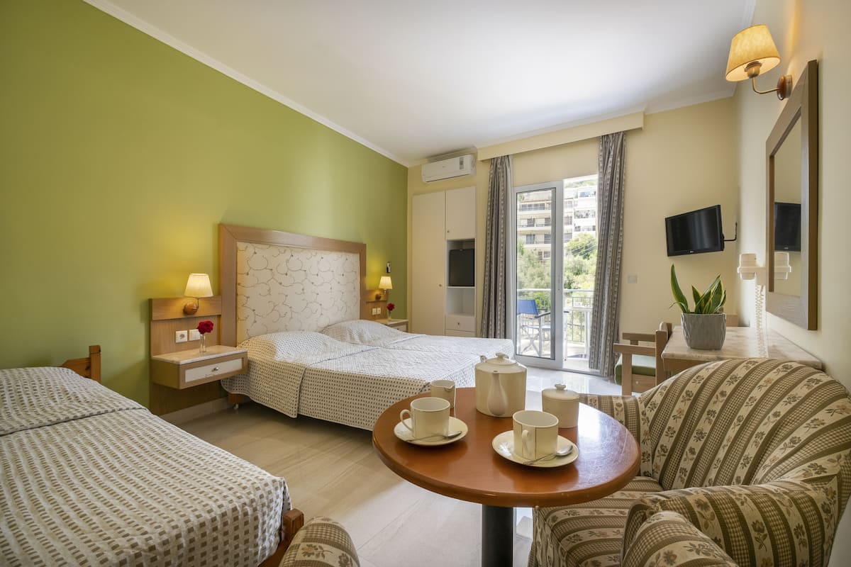 A hotel room with two beds, a dining table with cups, a sofa, a TV, and a window with curtains.