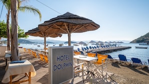 Ett område vid stranden med bord och stolar där man kan äta, en skylt med texten ”HOTEL DOLPHIN” och utsikt över båtar på vattnet.