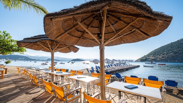En restaurang vid stranden med uteservering, parasoller med halmtak och utsikt över havet och bergen.