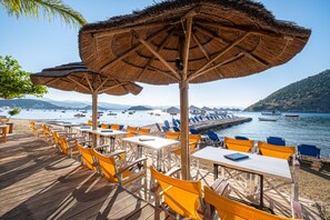 Un restaurante frente a la playa con terraza, sombrillas de paja y vistas al mar y a las montañas.