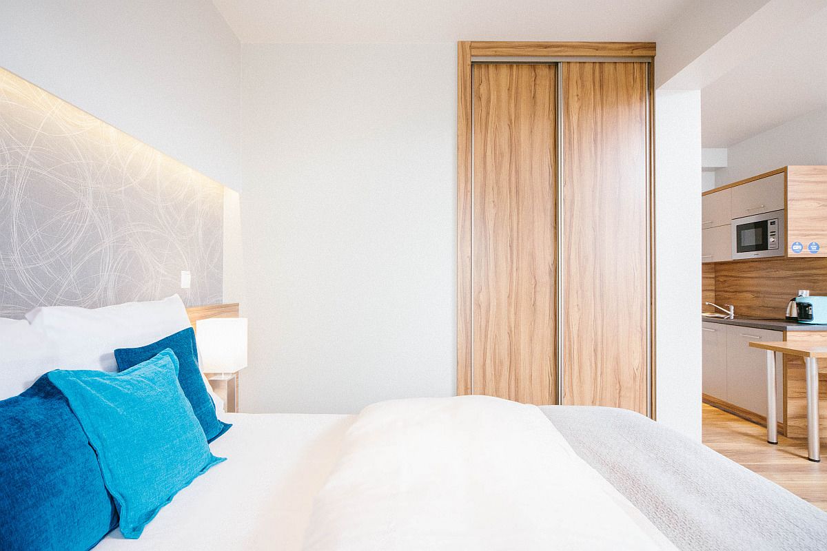A modern hotel room with a large bed, a wooden wardrobe, a kitchenette, and a nightstand with a lamp.